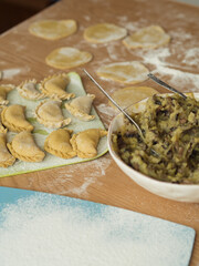 Preparing dumplings: A vertical close-up of Ukrainian varenyky on a cutting board, arranged in three rows. Dough circles are visible, with flour scattered across the table, a plate with filling and an