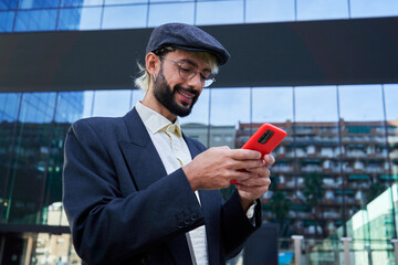 Young caucasian businessman using phone online texting, smiling, standing outside of a corporate district. Copy space image.