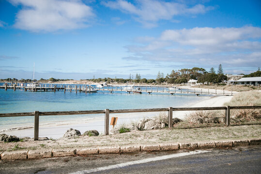 A road runs alongside a Thomson Bay, Rottnest Island, offering a view of the serene bay and jetty. The water reflects the sky above, creating a peaceful scene.