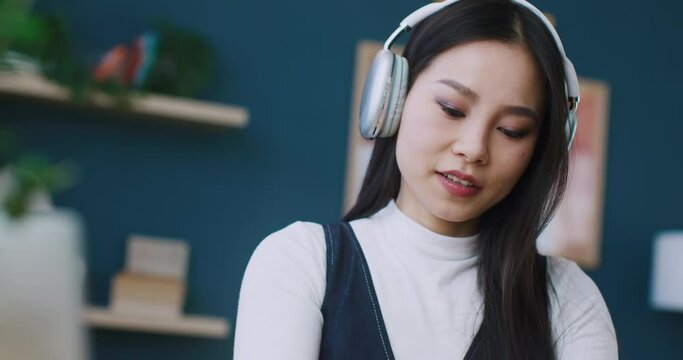 Pretty Asian woman wearing wireless headphones to enjoy some good music. Selecting her favourite audio on smartphone. Audio streaming from mobile phone. Having good feelings while listening media.