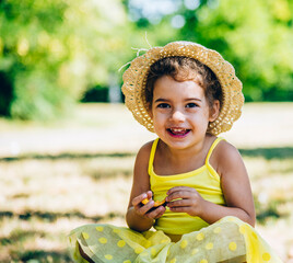 Cute little girl in yellow dress and summer hat enjoy eating fruit. Summertime child fun in countryside.