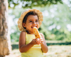 Cute happy little girl in yellow dress and hat eating fruit ice cream. Summer fun child playing in park. Summertime vibrant color background.