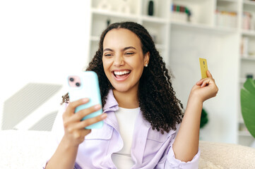 Joyful hispanic or brazilian woman, uses banking card and mobile phone to buy order prepay goods...