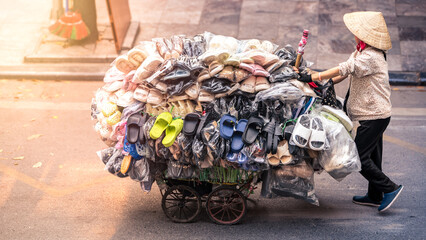 Street vendor wearing a straw hat, pushes a cart overflowing with shoes, Vietnam