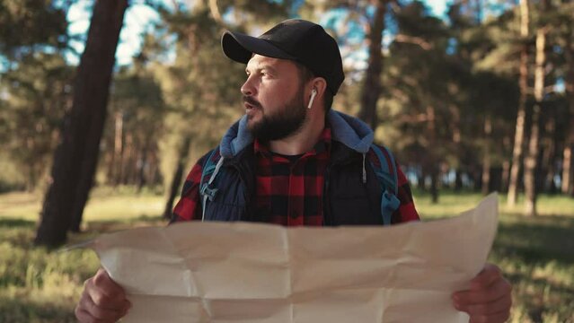 Active man hiking with map in forest. Tourist with carrying backpack hiking finding route way looking at paper map, walking on nature in forest park woodland. Tourism, trekking, hiking concept.
