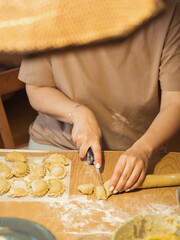 A vertical close-up captures a woman making dumplings, her face obscured by a straw lampshade, as she skillfully cuts dough with a knife.