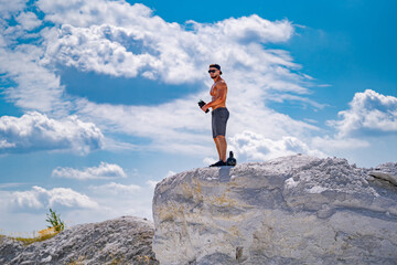 A man is standing on a rock with a camera in his hand. The sky is cloudy and the man is wearing an orange shirt