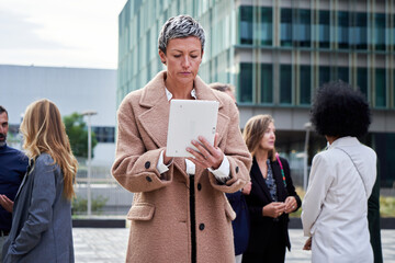Front view of a mature caucasian businesswoman with short hair working with a tablet outside office building, concentrated sending text messages or checking work projects. Copy space.