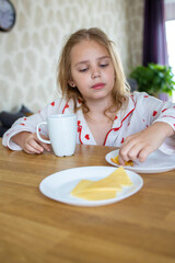 A pretty blonde girl in beautiful pajamas is sitting at a cozy table in the kitchen and having a delicious and healthy breakfast. The concept of proper and balanced nutrition.