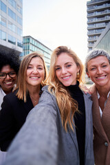 Cheerful vertical selfie of four multiracial businesswomen using phone to take self-photos together outside workplace looking at camera smiling. Copy space.