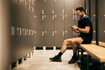 Side view of a smiling fitness man, sitting on the bench in the locker room, using a mobile phone,...