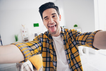 Photo of funky positive man wear checkered shirt stick out tacking selfie indoors house apartment...
