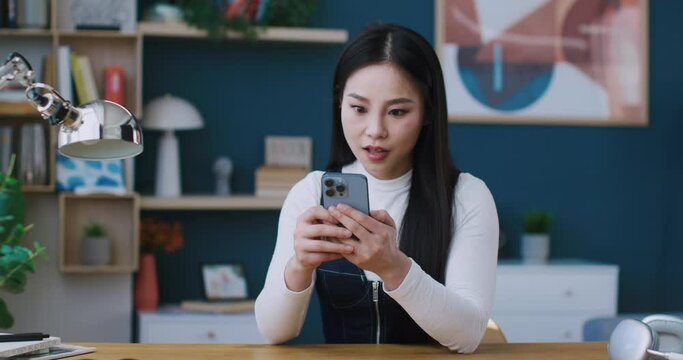 Adorable Chinese woman happy about getting positive notice on smartphone. Young woman covering her mouth in surprise while holding phone. Raising hand while celebrating achievement. Concept of success