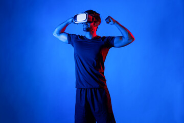 Happy man with VR goggle flexing muscle at neon light background. Healthy person with casual cloth...