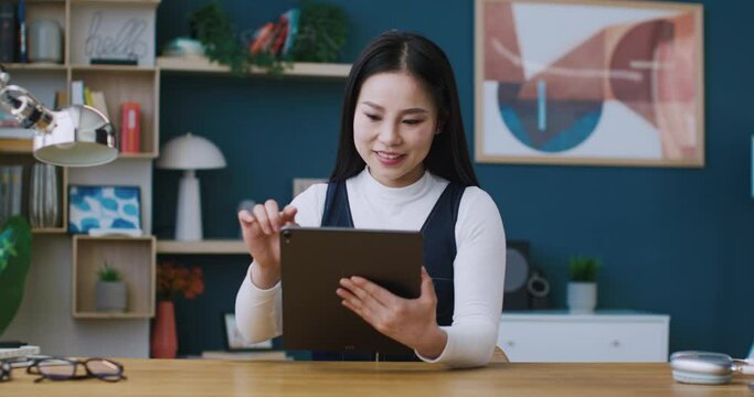 Young Asian student or worker searching information on tablet device for lesson or work. Joyfully smiling while using portative gadget. Browsing Internet with home network. Entering data on keyboard.