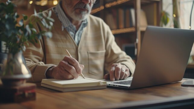 An elderly man studies online using laptop and takes notes in a notepad. Concept of the importance of learning for older people