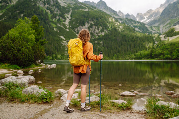 Naklejka premium Woman traveler with yellow hiking backpack and hiking stiks enjoys the scenery. Active lifestyle. Wanderlust.