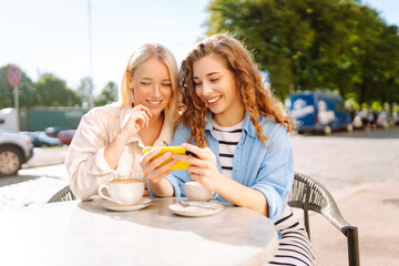 Two young women drinking coffee and using mobile phone while sitting at the summer cafe. Fashion, beauty, blogging, tourism.