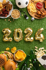 Get ready for Euro 2024 thrill: vertical top view of 2024 balloons, football snacks - chips, popcorn, fried chicken. Beer, soccer ball, whistle on grass background. Perfect for football passion