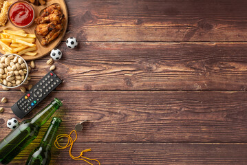 Top view of wooden table with football snacks including beer, fries, chicken wings, and remote,...