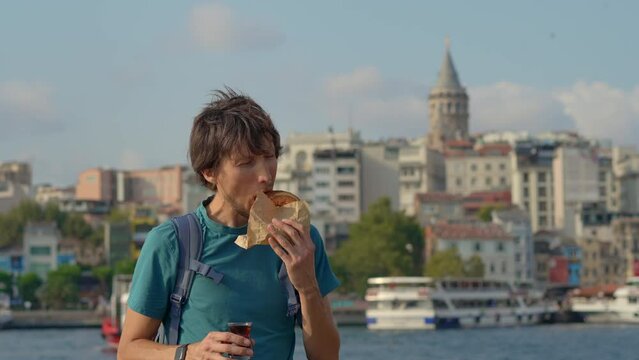 A young traveler relishes a bite of a crusty simit and sips Turkish tea at a pier with seagulls fluttering by and the iconic Galata Tower looming in the backdrop.