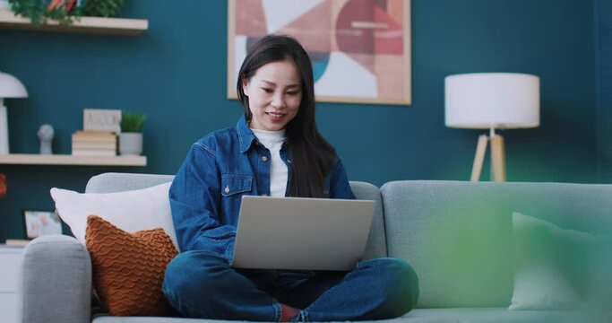 Camera view of cute Chinese woman using laptop for scrolling website or watching movie. Spending her free time with pleasure. Relaxation at home. Positively smiling while looking at screen.