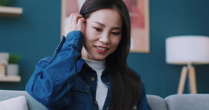 Beautiful Asian girl spending time on laptop while sitting on sofa in her cosy room. Fixing hair behind ear. Joyfully smiling while surfing websites or watching interesting movie on technology device.