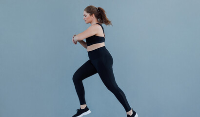 Side view of plus size female running against a grey wall wearing black fitness attire. Young woman...