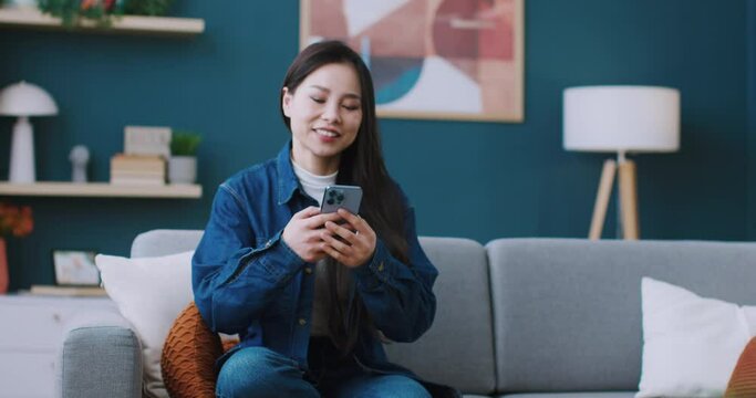 Pretty Asian woman walking to sofa and comfortably sitting. Actively messaging on phone with her friends or boyfriend. Smiling with joy during remote communication. Concept of using digital gadget.