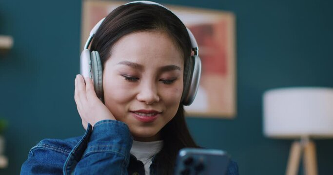 Happy Chinese woman with headphones listening music. Turning on favourite playlist on smartphone. Getting satisfied from good feelings after hearing amazing track. Moving hand with rhythm.