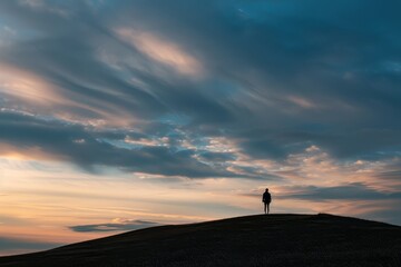 Fototapeta premium A person stands on a hilltop, looking out under a cloudy sky, A lone figure standing on a hilltop, watching the evening descend with a sense of peace and contemplation