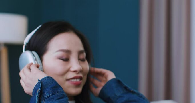 Portrait of Asian woman wearing wireless headphones on her head. Pretty girl with brunette hair closing eyes while starting to listen her favourite musical group or playlist. Tranquil atmosphere.