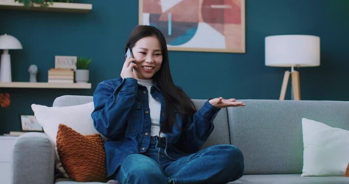 Happy Asian woman in jeans outlook using her cell phone while resting on soft couch at home. Cute girl joyfully smiling during remote communication on digital gadget. Concept of online conversation.