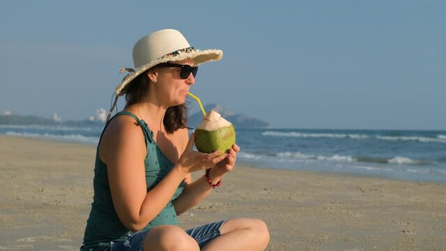 Cheerful young woman in straw hat drinking coconut water on the beach in the morning. Travel and vacation concept