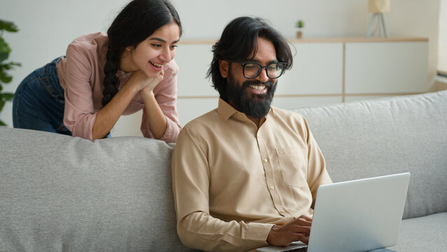 Arabian Indian couple on couch sofa use computer laptop shopping online internet store excited multiracial wife husband happy excited customers woman and man search goods purchase on website at home