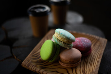 Multicolored macaroons in a wooden plate on the background of two cups of coffee in dark colors