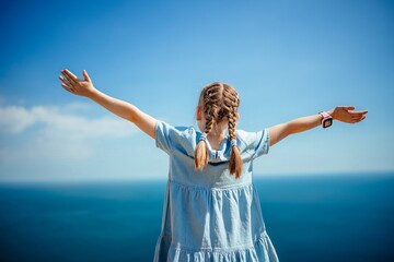 A young girl with pigtails is standing on a beach, looking out at the ocean. She is wearing a blue...