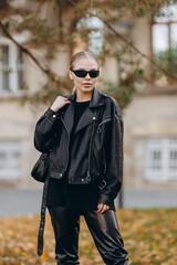 The modern woman with black sunglasses on her eyes is looking away from the camera and is wearing a black basic t-shirt, black leather jacket and black leather pants, holding a black bag