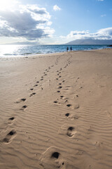 Pair of footsteps leading up to a couple of people walking towards the Pacific Ocean at Kamaole Beach Park II, golden sandy beach at sunset, Kihei, Maui, Hawaii
