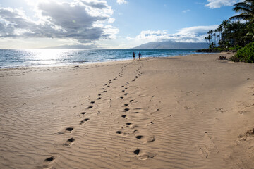 Pair of footsteps leading up to a couple of people walking towards the Pacific Ocean at Kamaole Beach Park II, golden sandy beach at sunset, Kihei, Maui, Hawaii
