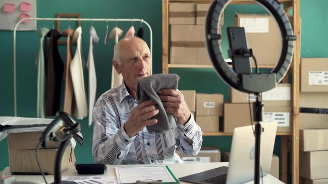 Blogger SME business old man using mobile phone and video call for sell clothes, packing product at home office, entrepreneur in e-commerce