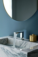 A closeup shot of the faucet, with water flowing from it in slow motion against a backdrop of blue walls and marble countertops. The mirror above reflects an atmosphere of luxury interior design.