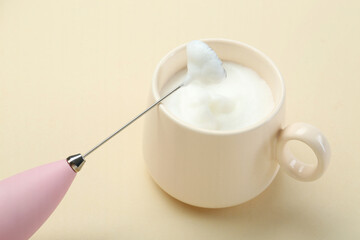 Mini mixer (milk frother) and cup of whipped milk on beige background, closeup
