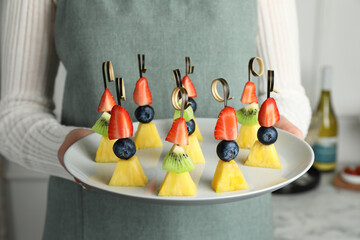 Woman holding plate of tasty canapes with pineapple, kiwi and berries indoors, closeup