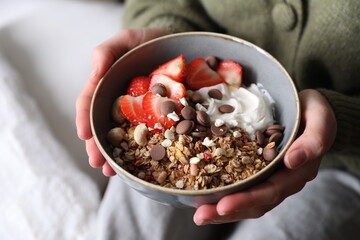 Woman holding bowl of tasty granola with chocolate chips, strawberries and yogurt indoors, closeup