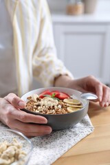 Woman eating tasty granola with banana, cashew and strawberries at wooden table indoors, closeup