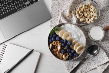 Delicious granola in bowl, stationery and laptop on white wooden table, flat lay