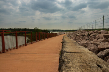 The path leads over a dam, granting a serene view of the water to the left and an expansive landscape in the distance. It's a picturesque spring day at an urban park in the Texas Hill Country.