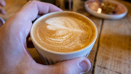 Close up of someone holding a hot, delicious cup of golden flat white coffee against a rustic wooden table in local cafe