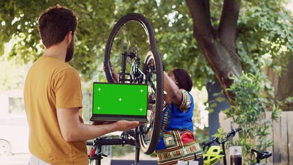 Young man with laptop displaying greenscreen as african american lady attaches wheel to bicycle...